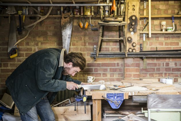 Hombre trabajando de pie en un banco de trabajo en un taller de carpintería, utilizando sierra en pieza de madera.