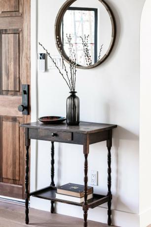 Uma mesa lateral com um vaso preto de mudas de salgueiro e um espelho redondo e porta de madeira