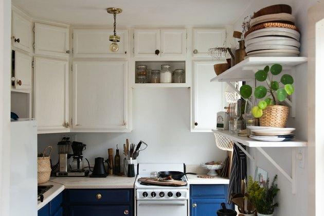 Cozinha eclética com armários suspensos brancos, armários inferiores azuis, fogão do tamanho de um apartamento, prateleiras abertas, pratos, plantas.