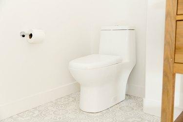 Un inodoro blanco en un baño con paredes blancas y piso de baldosas blancas y grises. Hay un rollo de papel higiénico en la pared junto al inodoro, colgado de un simple soporte cromado.