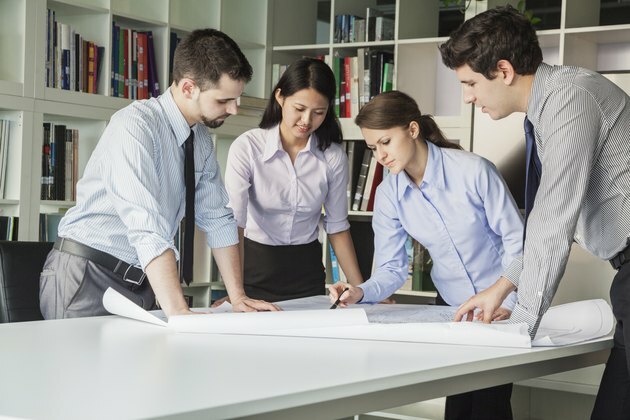 Neljä arkkitehtejä seisoo ja suunnittelee pöydän ympärillä katsellessaan suunnitelmaa