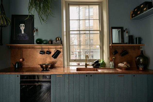 Rustieke kleuren in de keuken met gedempte groenblauw kasten, koperen toonbanken en backsplash, open planken, crème bekleding op raam.