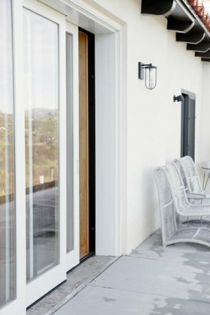 Perete alb cu ușă glisantă din sticlă, parțial deschis. O aplice de metal negru este pe peretele de lângă uşă. Două scaune de frânghie albă sunt mai jos, de-a lungul peretelui. Terenul este beton.