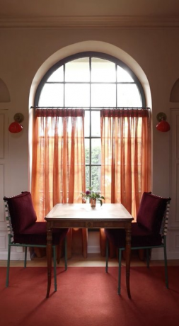 Tafel en stoelen, bordeauxrood tapijt, oranje gordijnen.