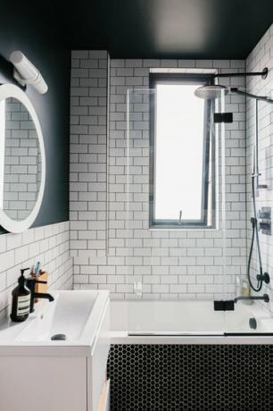 liten hvit vask, rundt speil med overlys, svart sekskantflis på badekaret, hvit undergrunnsflis på veggen, dypt badekar med svart kran og dusjhode
