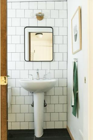 banheiro, parede grande de ladrilhos brancos do metrô, pia branca com torneira de prata, espelho retangular com detalhes em preto, toalha pendurada em verde