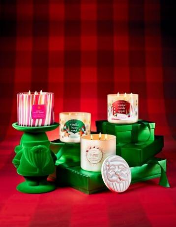 Rad zelených darčekov na pozadí červeného byvolieho šeku so štyrmi zobrazenými sviečkami Bath and Body Works s 3 knôtmi: Snowy Peach Berry, Vanilla Bean Noel, The Perfect Christmas a Winter Candy Apple