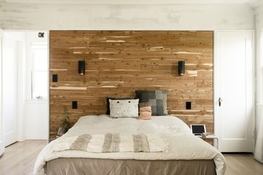 Uma cama com roupa de cama e travesseiros neutros multicoloridos. Uma parede de sotaque de madeira atrás com arandelas.