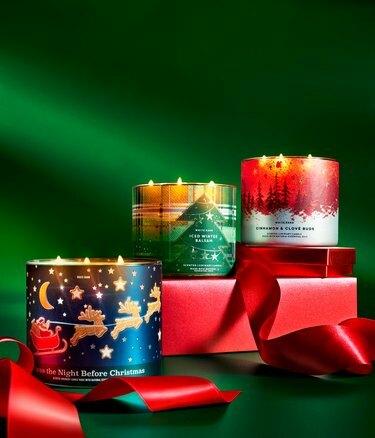 Hromada červených darčekov na zelenom pozadí s tromi vystavenými sviečkami Bath and Body Works s 3 knôtmi: Twas the Night Before Christmas, Iced Winter Balsam a Cinnamon and Clove Buds