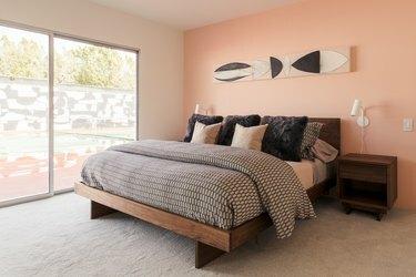 Quarto de parede rosa com roupa de cama preta e neutra. Uma mesa de cabeceira de madeira, arandelas de parede e pintura abstrata. Uma grande janela de imagem.