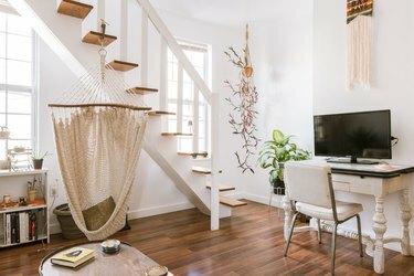 Uma mesa de perna de eixo branco com um computador de mesa. Planta de casa e decoração de arte têxtil. Uma escada de madeira e piso de madeira.