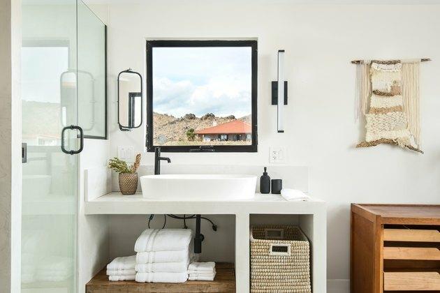 hvit forfengelighet av baderom, hvit keramisk vask med svart kran, åpent vindu, hengende veggteppe, oppbevaringskurv, hvite håndklær, treoppbevaring, glassdør
