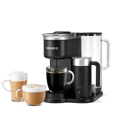 Naujausias Keurig alaus darykla, K-Cafe SMART, yra juodos ir sidabrinės spalvos kavos virimo aparatas su juodos ir sidabrinės spalvos pieno broliu. Skaidrus kavos puodelis gaudo paruoštą kavą, o kiti du skaidrūs puodeliai, užpildyti latte, yra šone.
