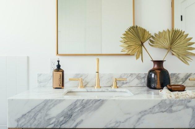 marmor forfengelighet benkeplate med keramisk undervanns vask, gullkran og håndtak, brun såpedispenser, vase med papirblomster, rektangulært speil med gullpynt