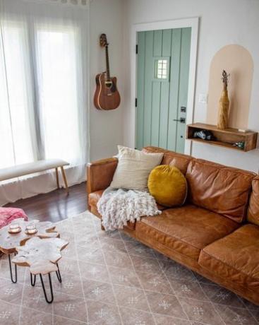 Un divano in pelle marrone con cuscini e una coperta su un tappeto rosa chiaro a motivi geometrici. Una mensola e una chitarra sono appese al muro e la porta è di un verde chiaro.