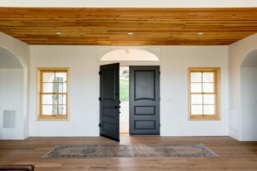 Hol wejściowy domu w stylu hiszpańskim z podwójnymi drzwiami z ciemnego drewna, z których jedne są otwarte, na białej ścianie. Dwa okna z ramami z jasnego drewna po obu stronach drzwi. Podłoga i sufit są wykonane z drewna. Przed drzwiami jest dywan.