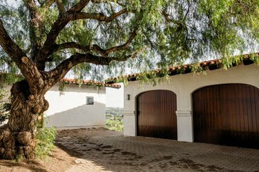 Прилаз од цигле који води до беле гараже у шпанском стилу са двоја дрвена врата. Храст поред прилаза.