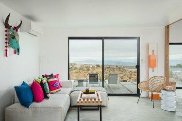 रंगीन तकिए, बड़ी खिड़कियों और लकड़ी के लहजे के साथ धूसर सोफे के साथ न्यूनतम रहने का कमरा