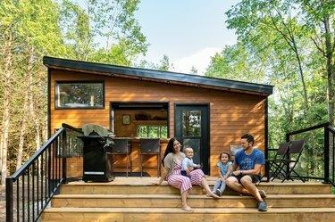 Scatto all'aperto di una moderna casa rustica con pannelli in legno con terrazza all'aperto con genitori e bambini contro alberi e cielo blu