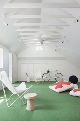 Witte zolder speelkamer met groen astro gras, witte canvas vlinderstoelen, oranje en witte vloerkussens, fiets, bijzettafel, plafondventilator.