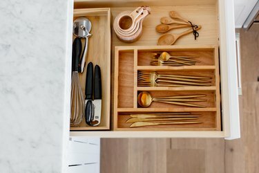 Een open lade met een houten bestekopberger. Gouden bestek, maatbekers en lepels en andere gebruiksvoorwerpen zichtbaar binnenin. Marmeren aanrecht boven de lade.