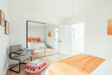 नारंगी तकिए के साथ सफेद बिस्तर के साथ बेडरूम, हल्की लकड़ी का फर्श, दर्पण वाली अलमारी और ग्रे आर्मचेयर