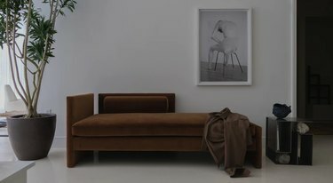 Una sala de estar blanca con una planta en maceta, una mesa lateral geométrica y una escultura, y una tumbona marrón con una camisa marrón esparcida sobre ella se encuentra contra la pared debajo de una fotografía en blanco y negro.