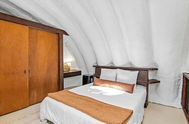 Белая кровать с коричневыми акцентами рядом с деревянным шкафом под белым сводчатым потолком