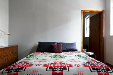 Bed met grijze kussens, rood geruit accentkussen, sprei in het zuidwesten en een houten dressoir met een witte lamp.