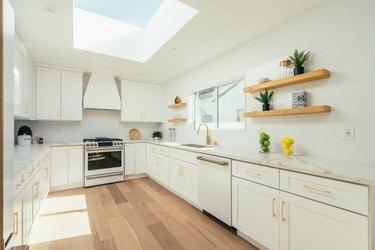 Baltos spintelės su auksinėmis rankenomis, stoglangiu, medinėmis grindimis minimalistinėje virtuvėje