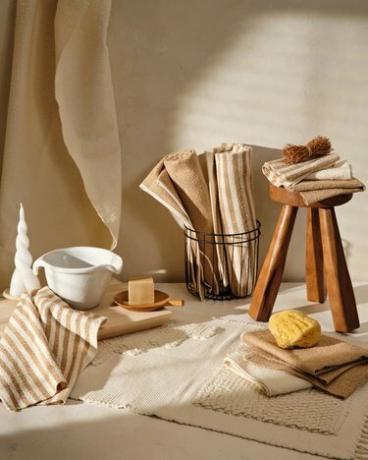 Beige-weiß gestreifte und einfarbige Handtücher werden in einem Badezimmer mit Holzhocker und -tablett, schwarzem Drahtkorb und weißem Keramikkrug gefaltet und gerollt