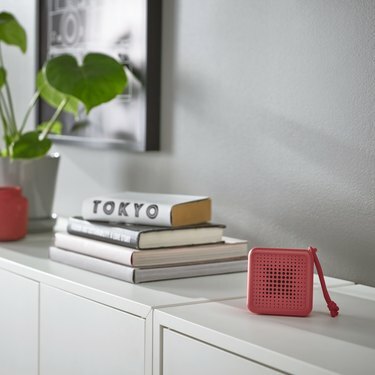 ИКЕА ВАППЕБИ црвени звучник лежи на белој конзоли, поред хрпе књига и биљке.