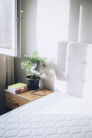 Witte matras naast bijzettafel met lamp en plant in witwandige kamer met witte gordijnen