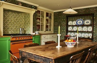 Maksimalistinen keittiö, jossa on oranssivalikoima, vihreät kaapit, maalaismainen ruokapöytä, astiateline, lasikaappi