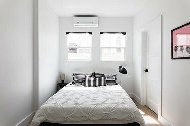 सफेद बिस्तर और काले लहजे के साथ एक न्यूनतम सफेद दीवार वाले बेडरूम में डक्टलेस मिनीस्प्लिट एयर कंडीशनर