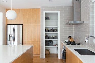 Keuken met houten kasten, ronde witte hanglamp, witte toonbanken, grijze tegels, houten vloeren. Een voorraadkast met grijze manden.